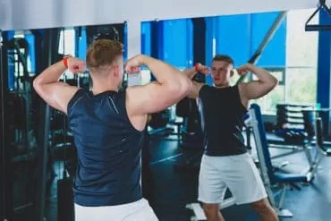 man in a gym doing a front double biceps pose in front of a mirror