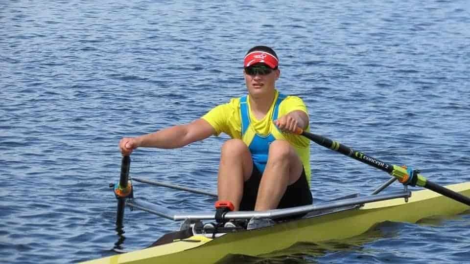 a male rower in a single scull, wearing blue and yellow suit and a red cap
