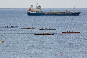 several rowing teams racing in a coastal rowing event and passing by a big vessel